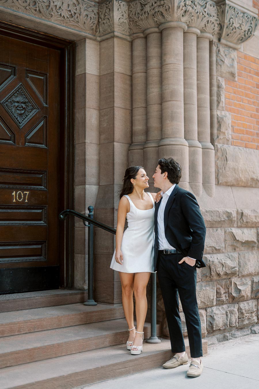 A couple in formal attire stands on stone steps outside a historic stone building, sharing a romantic moment in Ontario, Canada.