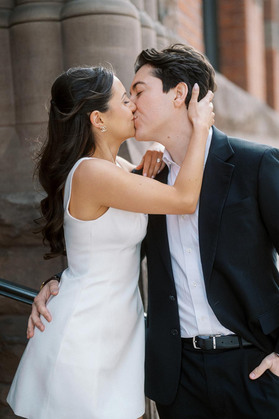 A couple in formal attire shares a kiss in front of stone columns and brick walls in downtown Ontario, Canada.
