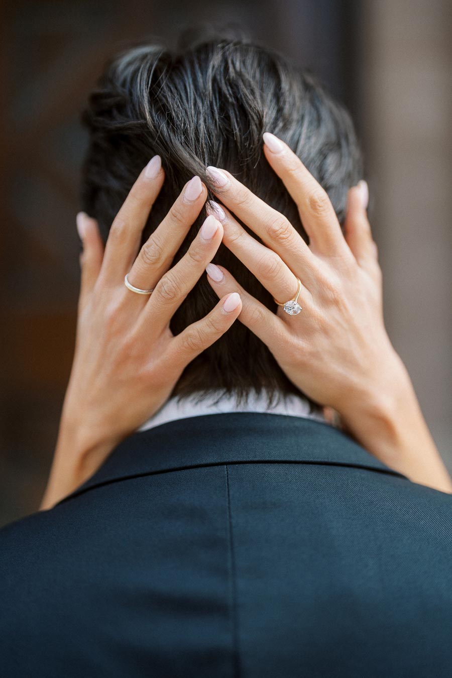 Close-up of a woman's hands with a diamond engagement ring resting on the back of a man's head, both dressed formally.