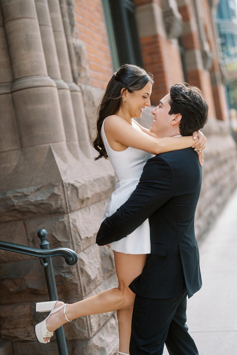 A couple embracing and smiling on a city sidewalk in front of a historic stone building, the woman in a white dress and the man in a dark suit.
