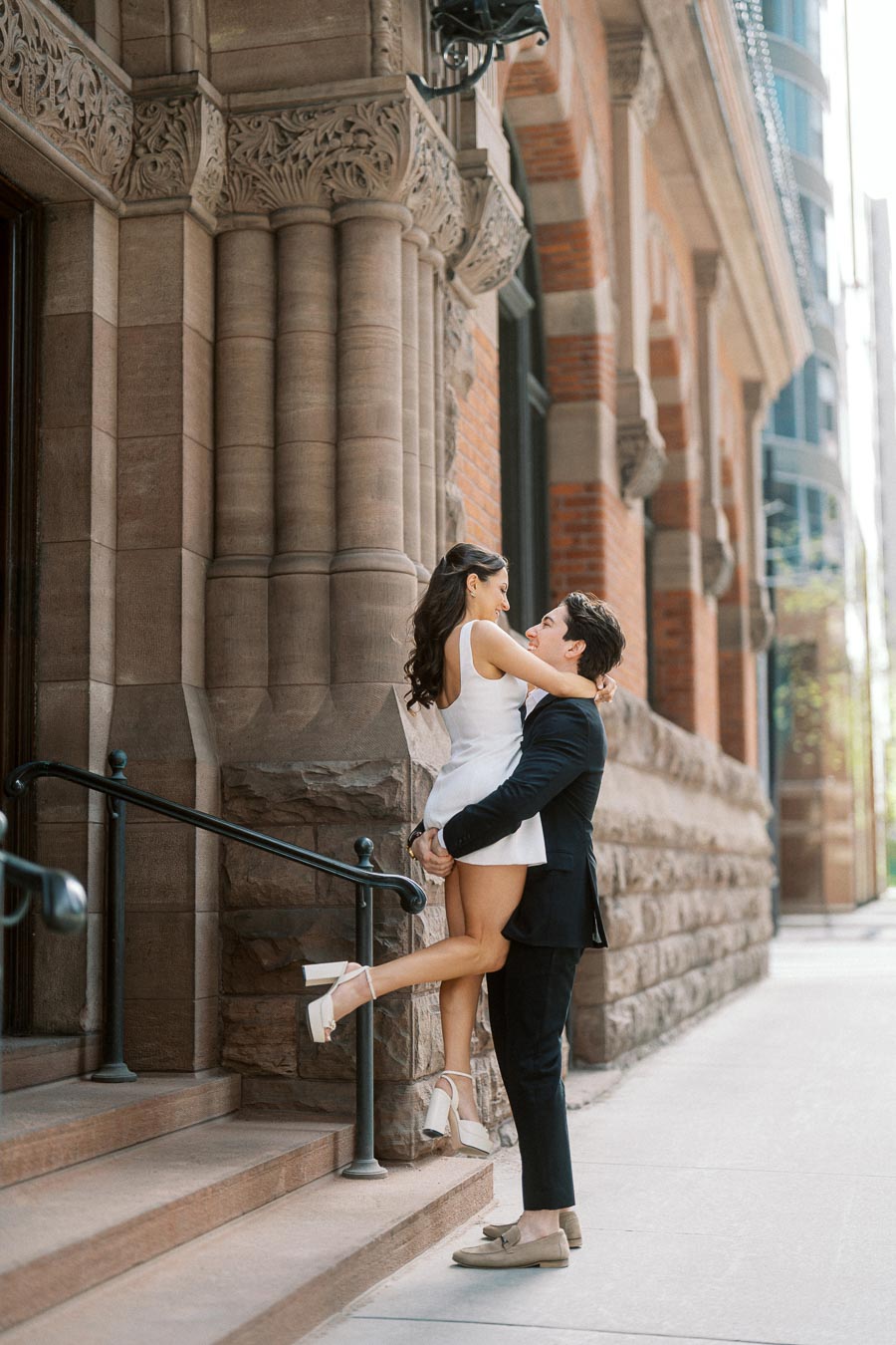 A man in a navy suit lifts a woman in a white dress on the steps of a historic stone building in downtown Ontario, Canada.