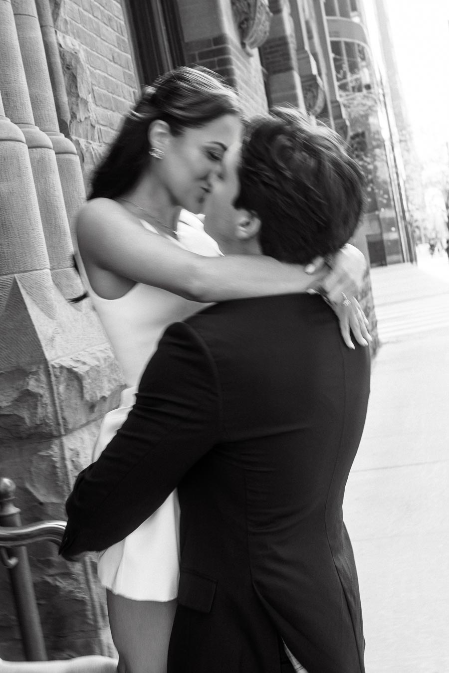 A couple embracing and sharing an intimate moment on a city sidewalk in front of historic stone buildings, black and white photo.
