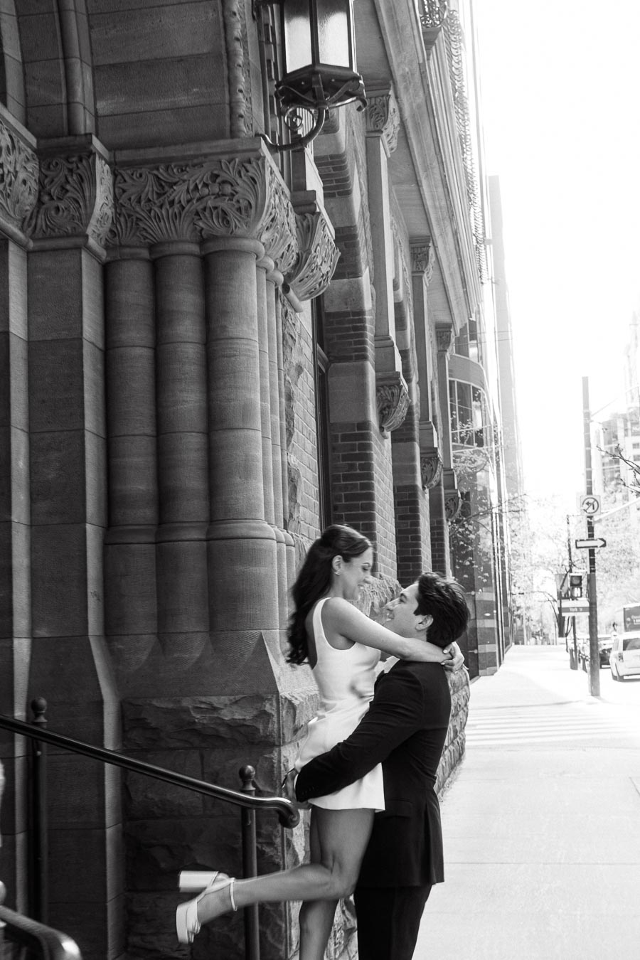 A man lifts a woman in a white dress in front of an ornate stone building on a city sidewalk, both smiling in an urban setting.