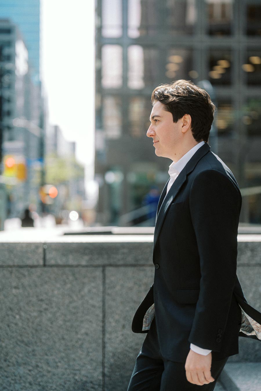 A man in a dark suit walks beside a stone wall with modern office towers and city streets in the background, Ontario, Canada.