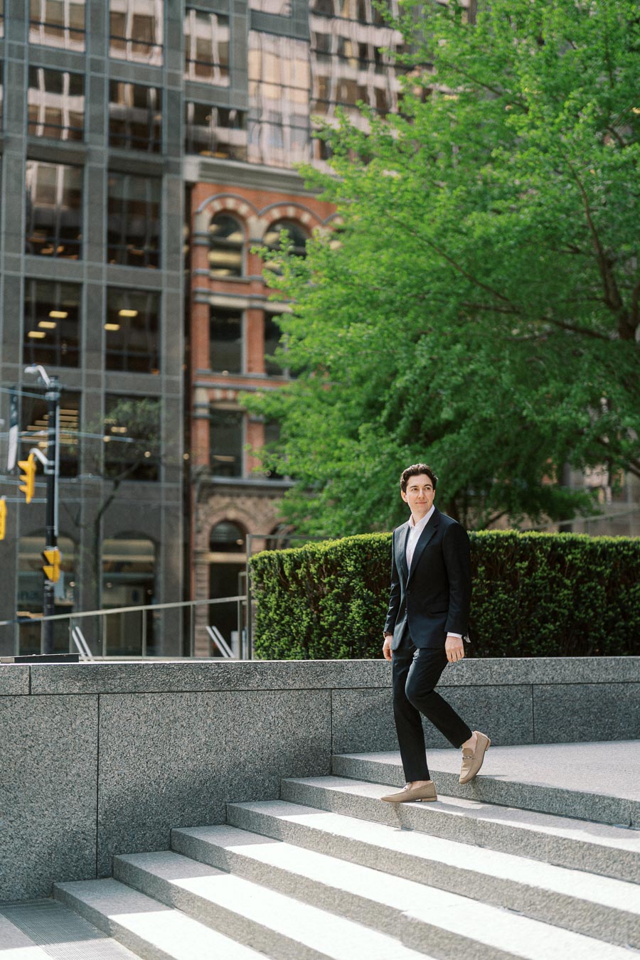 A man in a suit descends granite steps in a modern urban setting with glass office towers, historic stone buildings, and lush green trees.