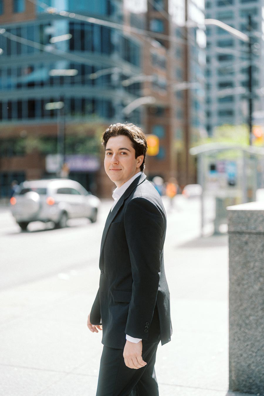 Man in a dark suit walking on a sunlit downtown street with modern glass and brick buildings in the background, cars passing by.