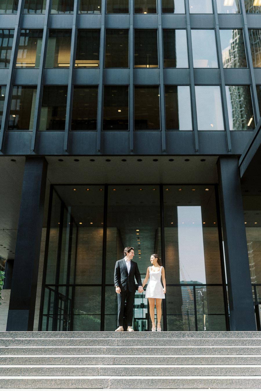 A couple stands hand in hand on city steps in front of a modern glass and stone office building, with urban reflections visible.