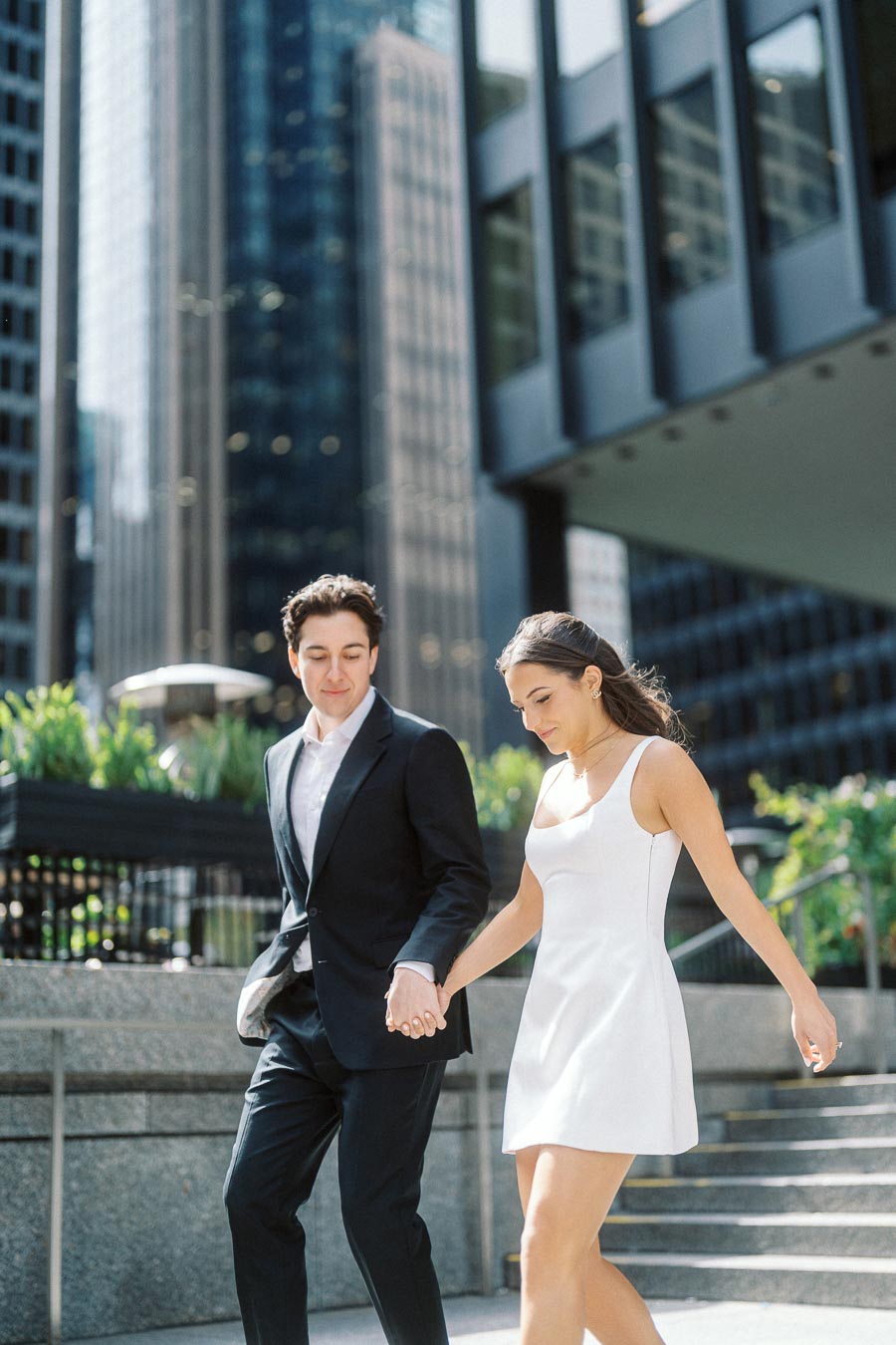 A couple holding hands walking down sunlit city steps with modern office towers and greenery in downtown Toronto.