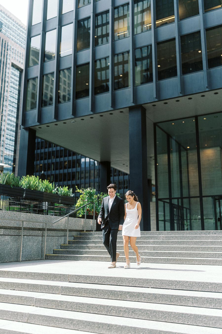 A couple dressed in formal attire walks down wide stone steps in a modern urban setting with office towers and glass buildings in the background.