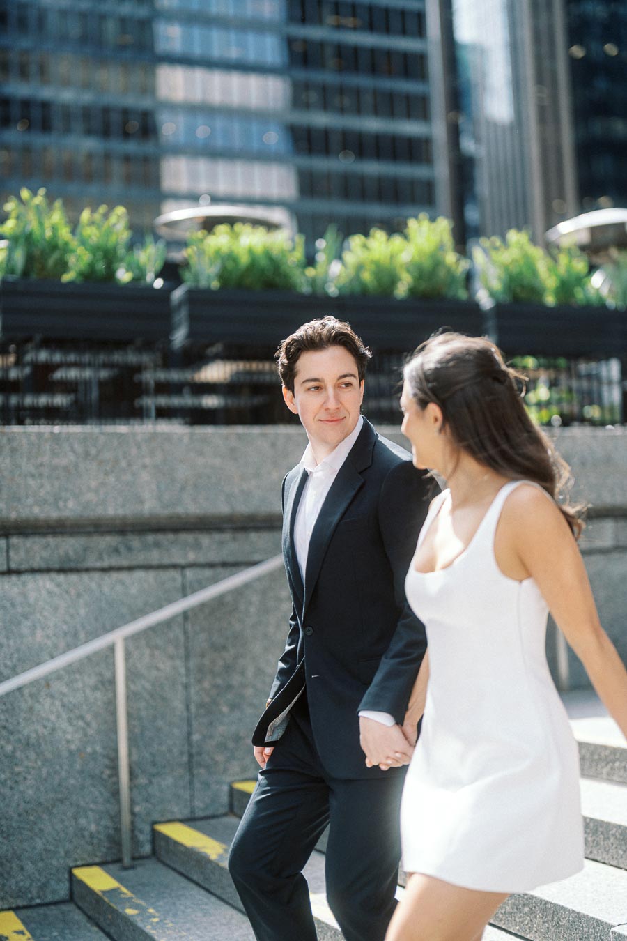 A couple dressed in formal attire walks hand in hand down outdoor city steps, with modern office buildings and greenery in the background.