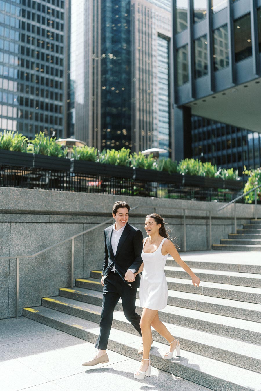 Couple holding hands and walking down wide stone steps in a downtown urban setting with tall office towers and greenery in the background.