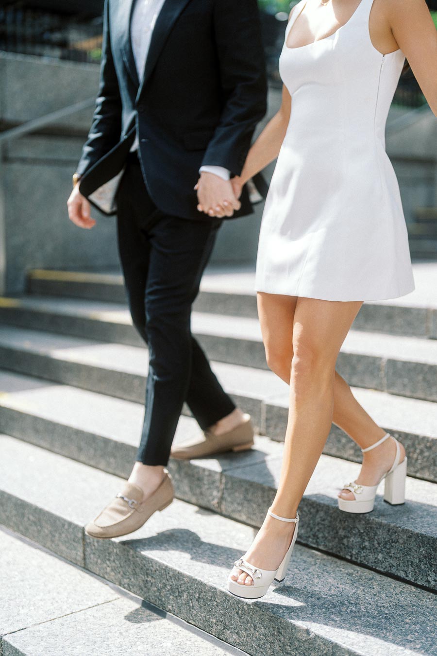 A couple dressed in formal wear holding hands and walking down sunlit stone steps in an urban setting, only lower bodies visible.