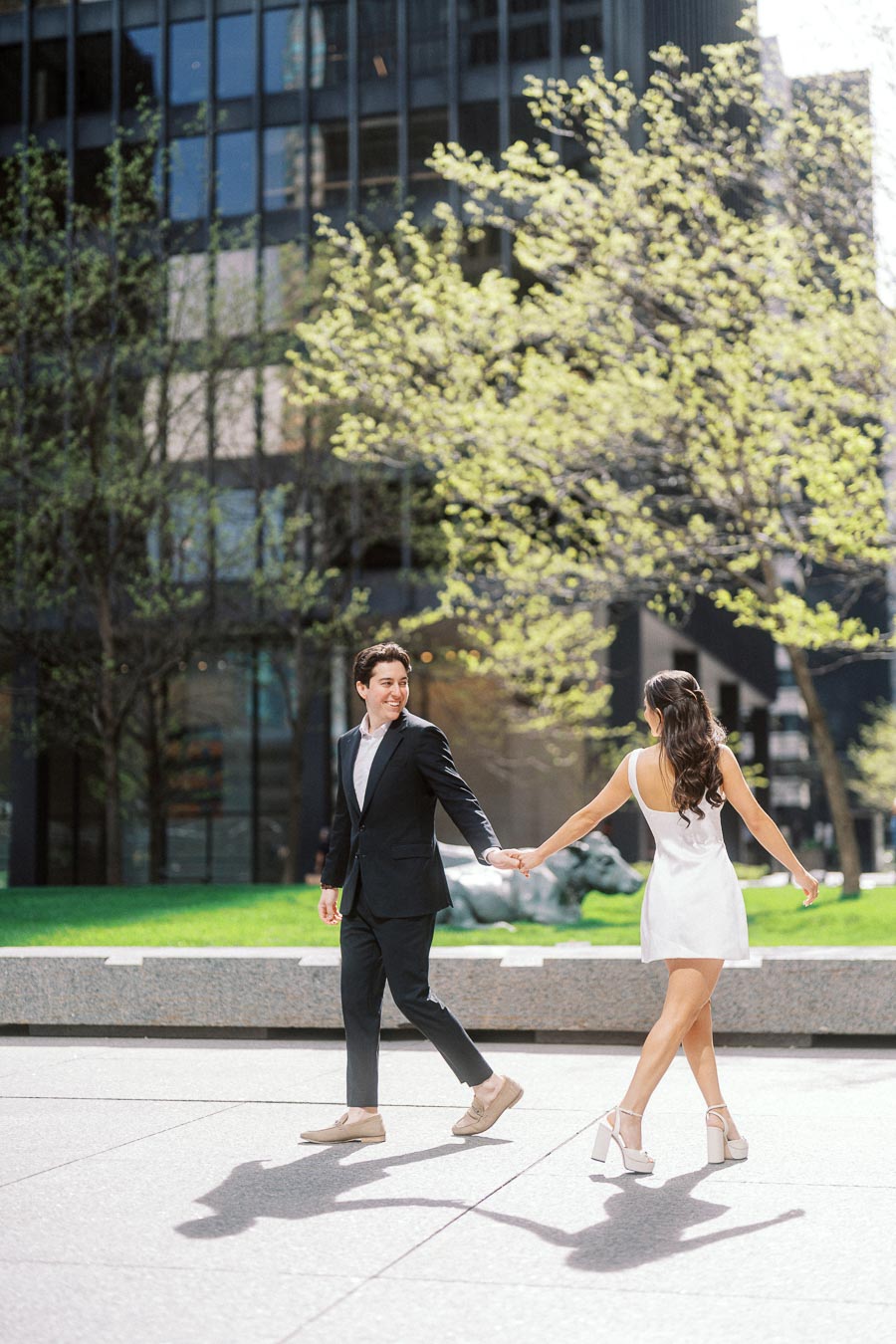 A couple in formal attire holding hands, walking on a sunlit city plaza with green trees and modern glass office buildings in the background.