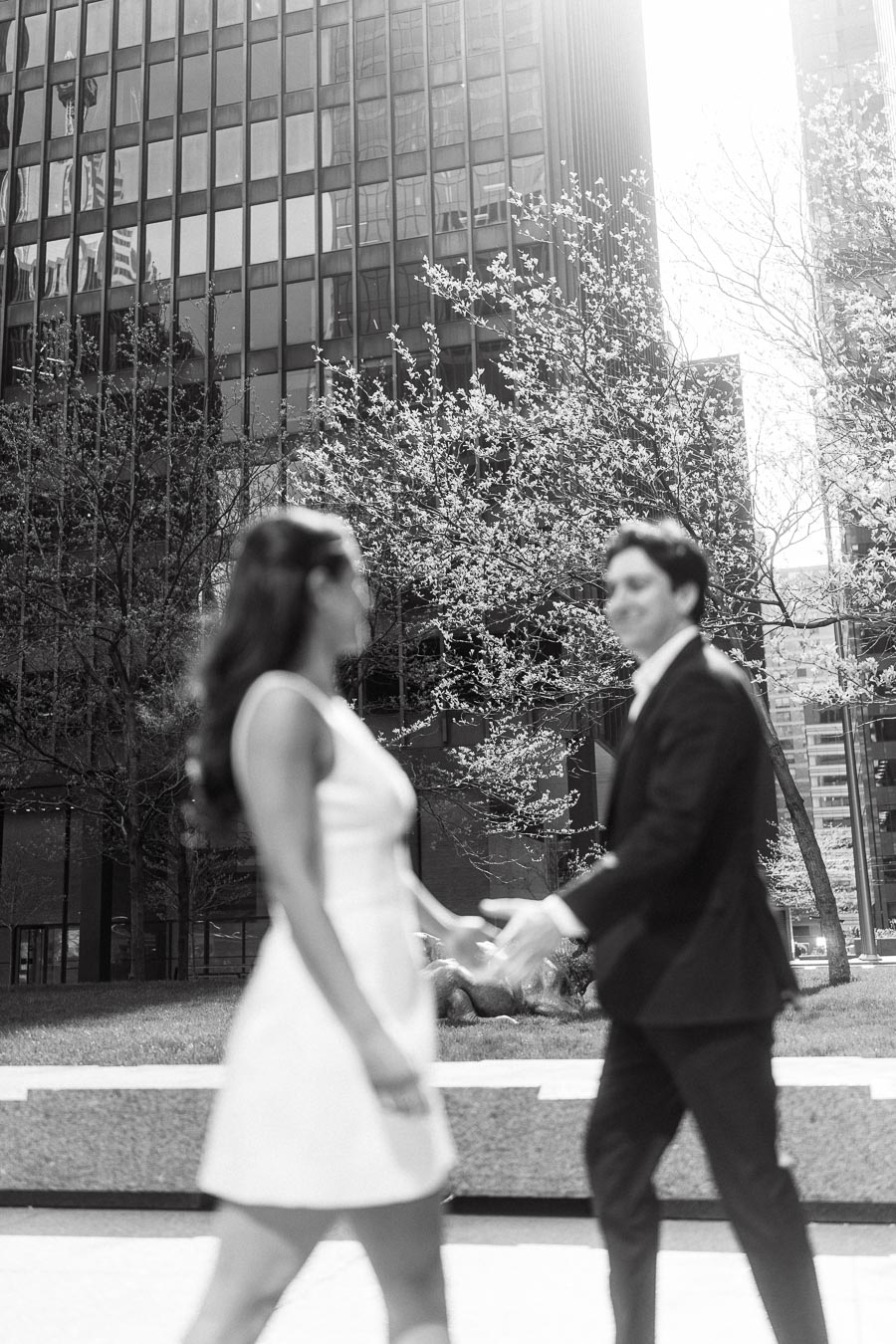 A blurred couple holding hands in front of glass office towers and blooming trees in a modern urban downtown setting, black and white photo.