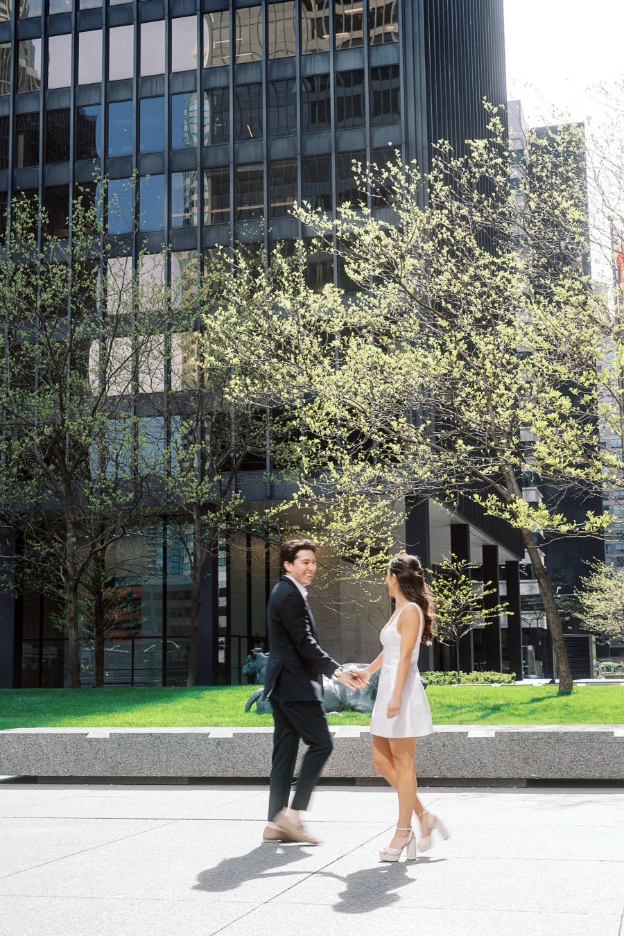 A couple dressed in modern attire hold hands and smile at each other on a city plaza surrounded by glass office towers and spring trees.