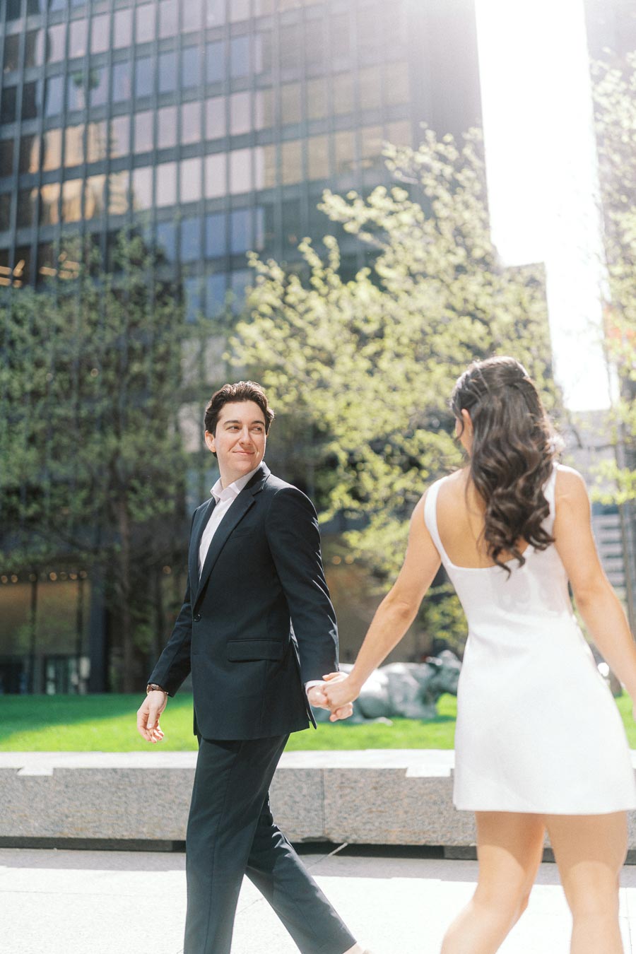 A couple holds hands and walks outside in downtown Toronto near glass office towers and green trees, sunlight shining through.