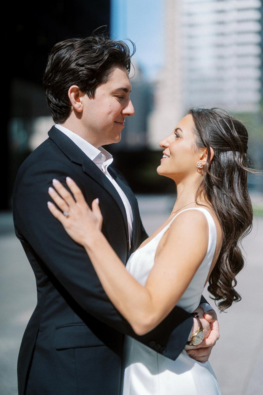 A couple dressed in formal attire smiles at each other while embracing outdoors in a sunlit urban setting with modern buildings.