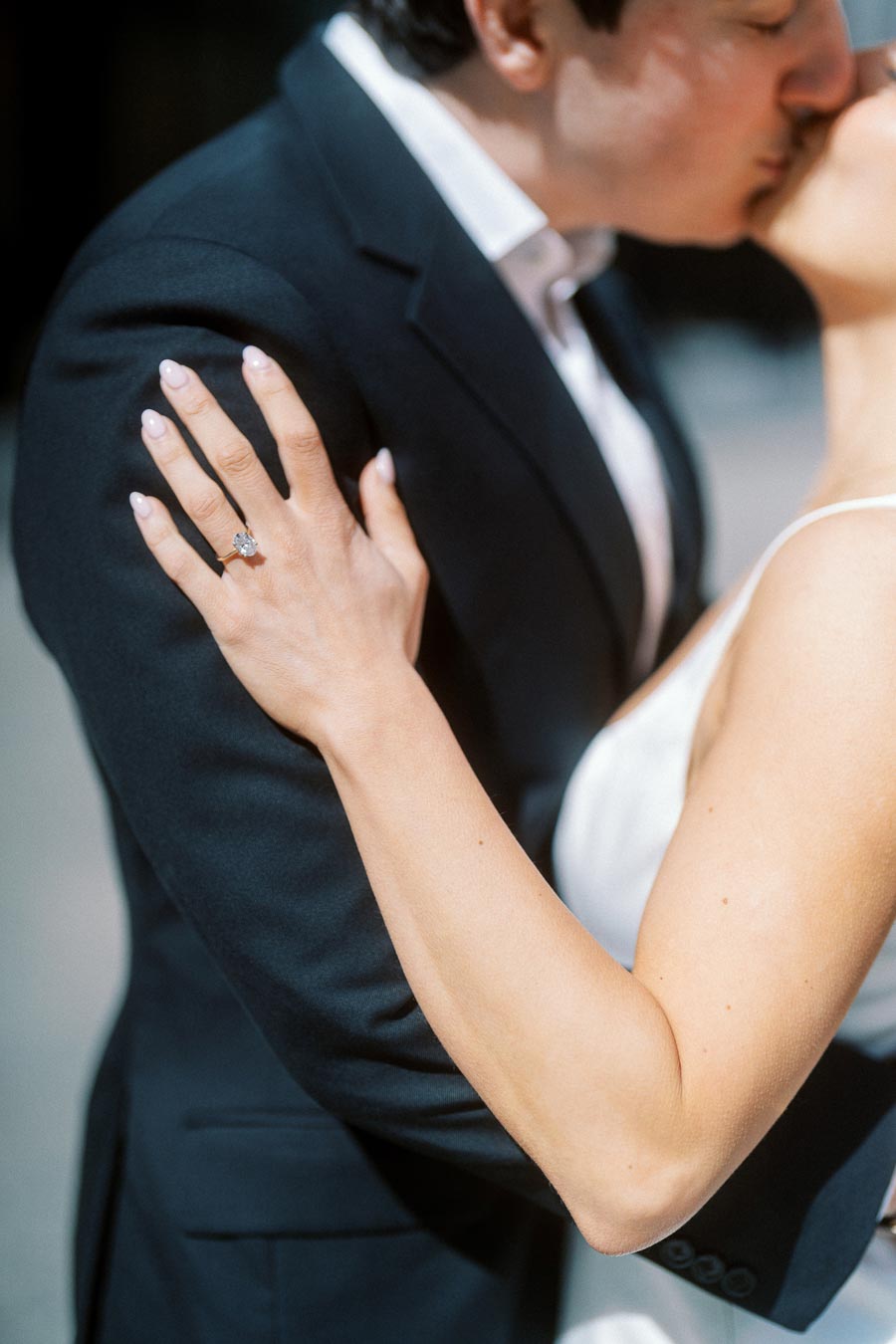 Close up of a couple kissing, woman in a white dress with hand on mans shoulder showing a diamond engagement ring, outdoors in soft sunlight.