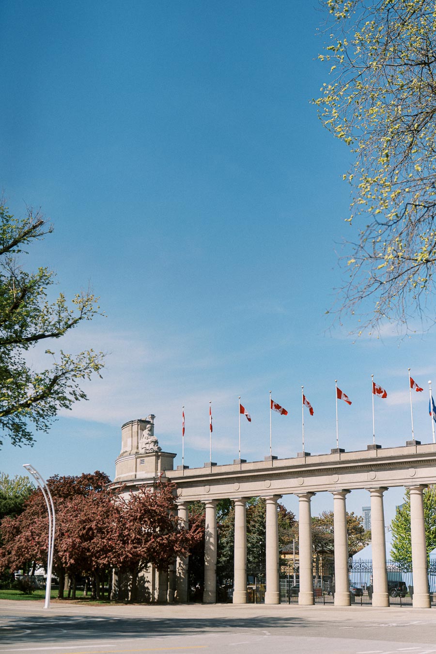 Princes' Gates at Exhibition Place in Toronto with Canadian flags atop, surrounded by spring trees and clear blue sky, photographed in May.