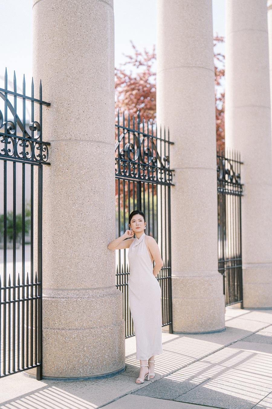 Bride-to-be in a white dress stands by the stone columns and black iron gates of Princes Gates at Toronto's Exhibition Place, with spring trees in the background.