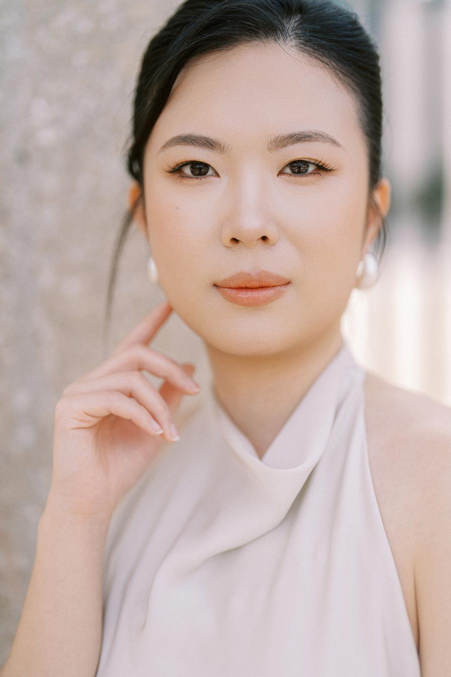 Close-up of an Asian bride-to-be with natural makeup, pearl earrings, and a white dress, gently touching her face during a Trillium Park engagement session in Toronto.