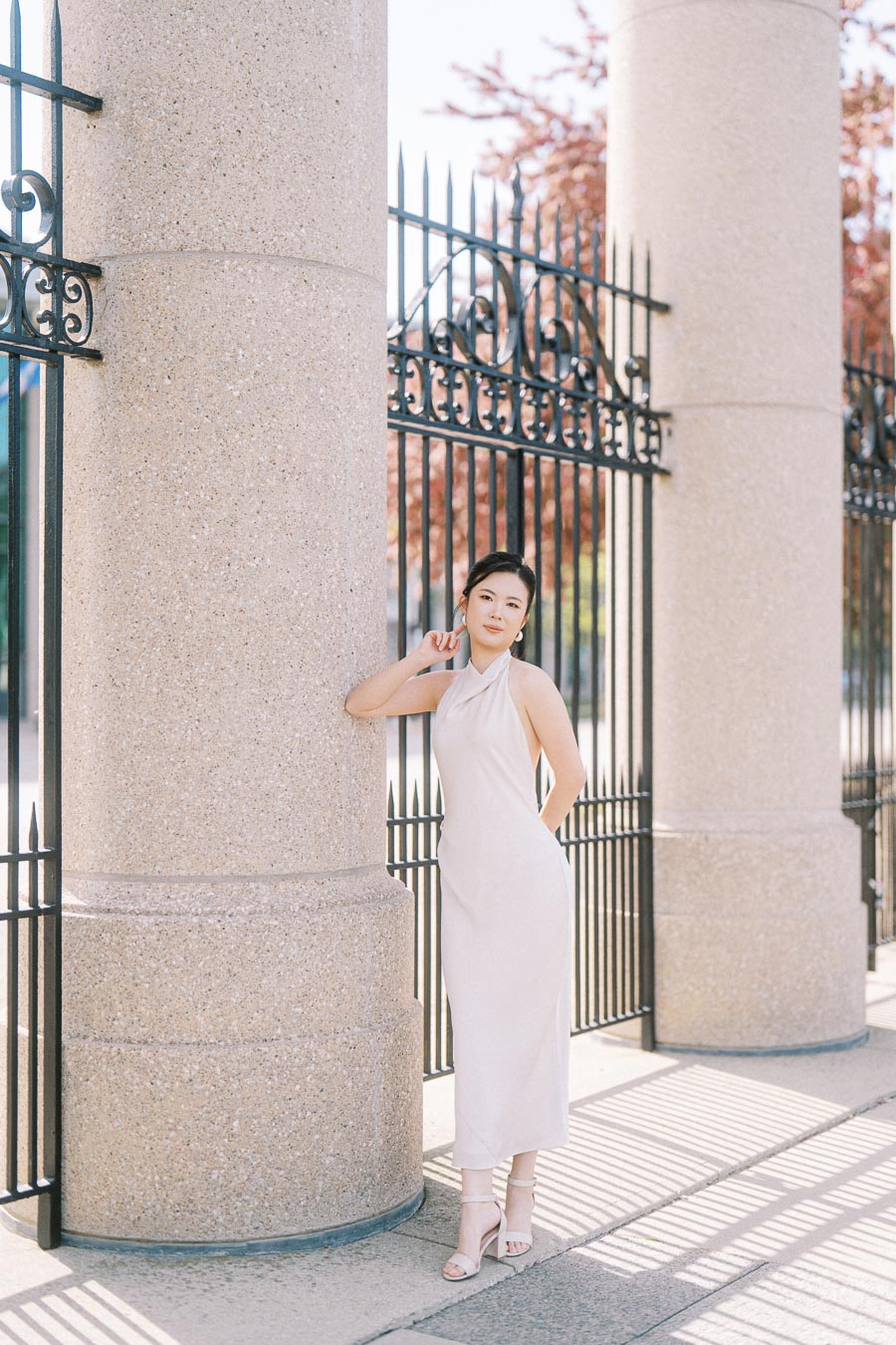Bride-to-be in a white dress leans against a large stone column at Princes Gates, Toronto, with ornate black iron fence and spring trees in the background.