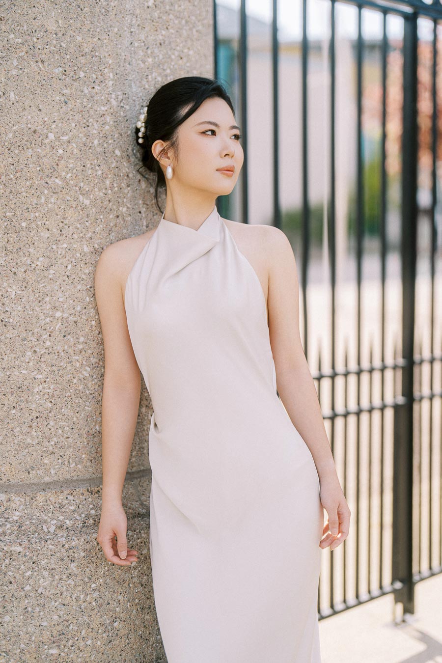 Bride-to-be in an elegant white dress leans against a stone pillar at Trillium Park, Toronto, with soft natural light and a black iron fence in the background.