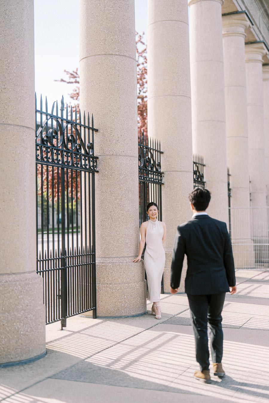 Asian engaged couple at Princes Gates, Toronto. Woman in a white dress leans against a column, smiling at her partner approaching in a black suit, with iron gates and soft sunlight.