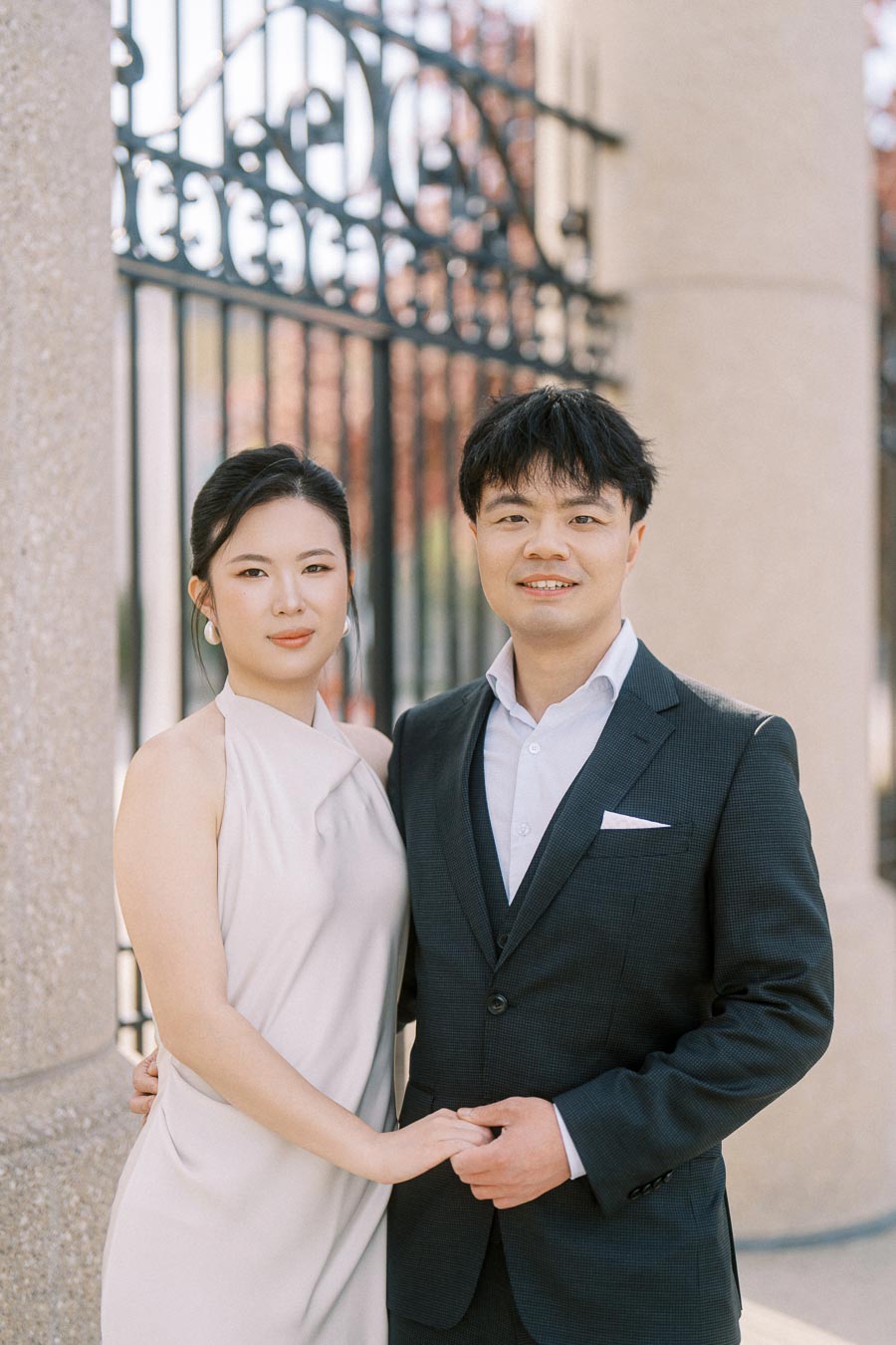 Engaged Asian couple standing close together, holding hands and smiling softly in front of the ornate Princes Gates at Trillium Park, Toronto.