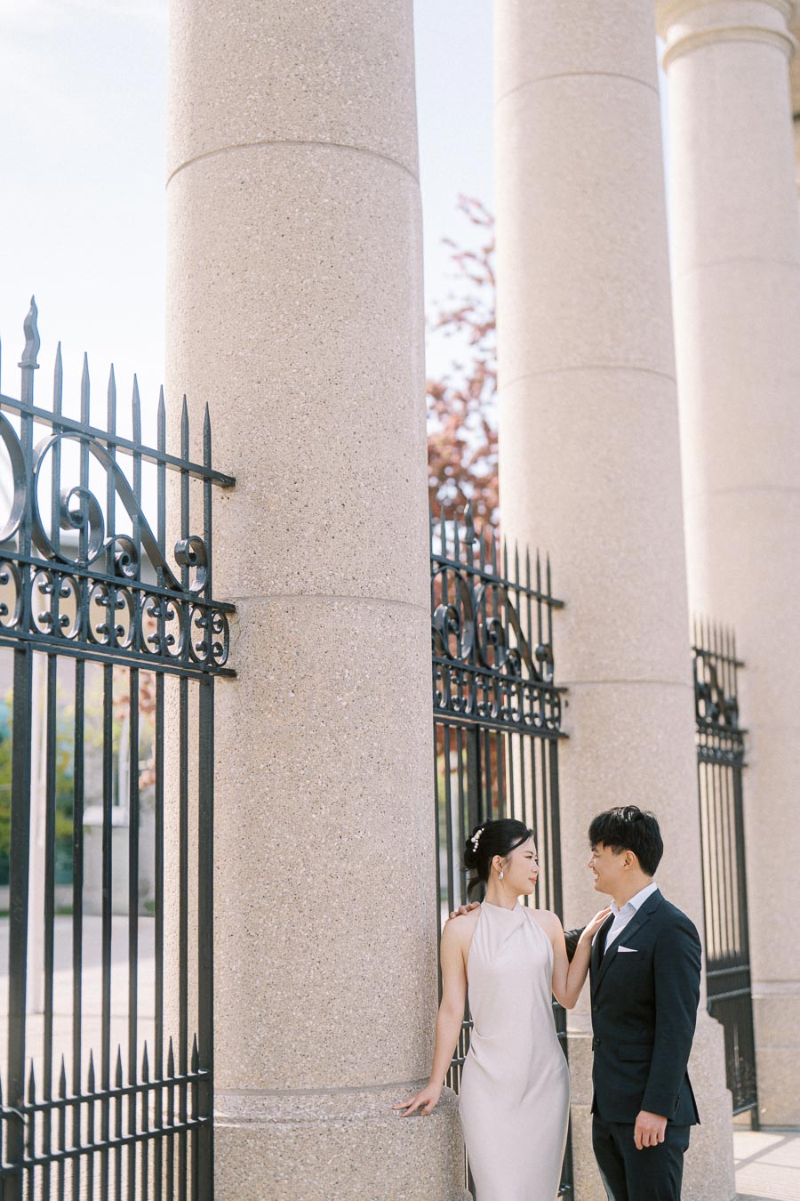 Asian engaged couple stands together by large stone columns and black iron gates at Princes Gates, Toronto, sharing a romantic gaze on a sunny spring day.