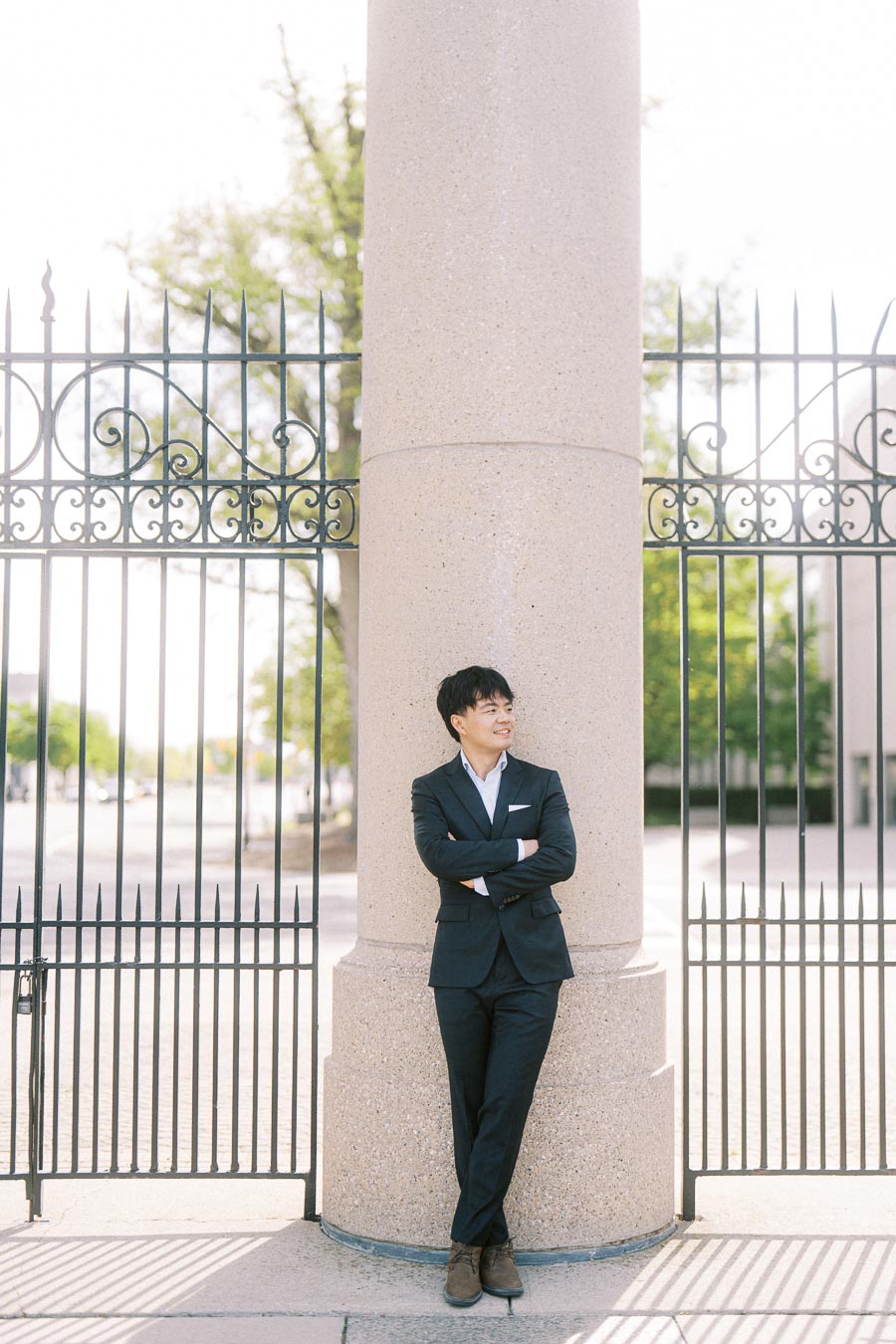 Asian groom-to-be in a dark suit leans against a stone pillar at Princes Gates in Toronto, with iron gates and trees in the background.