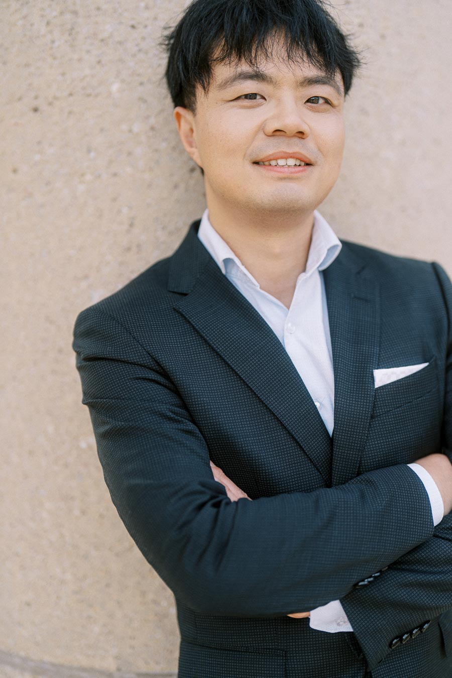 Asian groom-to-be in a black suit stands with arms crossed, leaning against a textured stone wall at Trillium Park, Toronto.