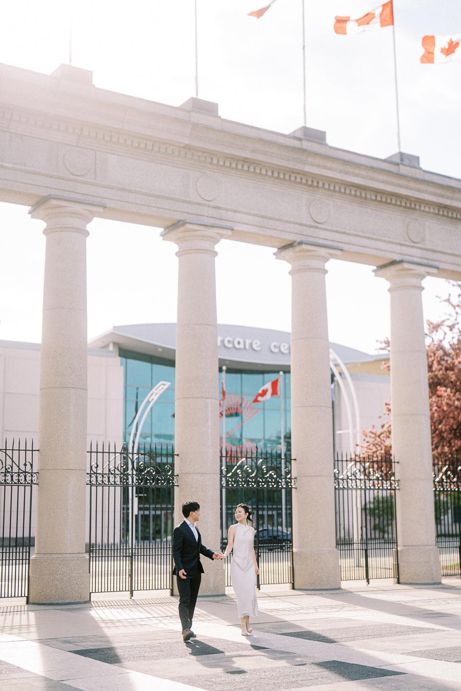 Engaged couple walking hand in hand beneath the Princes Gates at Exhibition Place, Toronto, with Canadian flags and spring sunlight in the background.