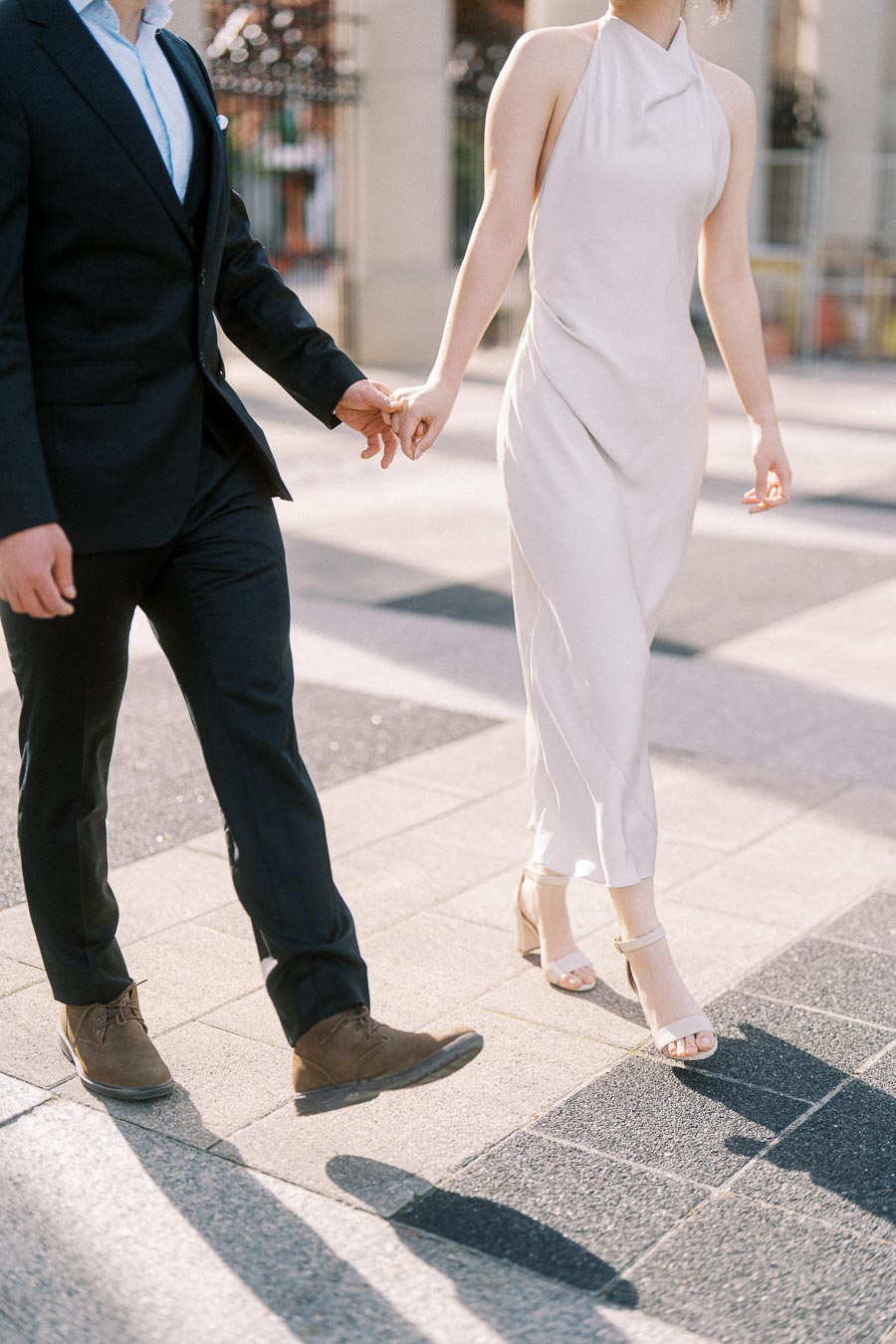 Asian engaged couple walking hand in hand on a sunlit paved path at Trillium Park, Toronto. The woman wears a white dress and the man a black suit, captured mid-step.
