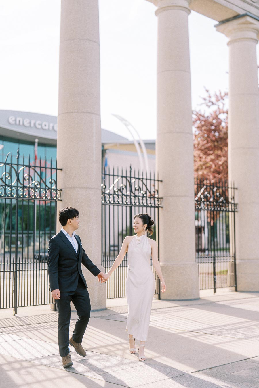 Asian engaged couple walking hand in hand in front of the stone columns and black iron gates of Princes' Gates at Exhibition Place, Toronto, on a sunny day.