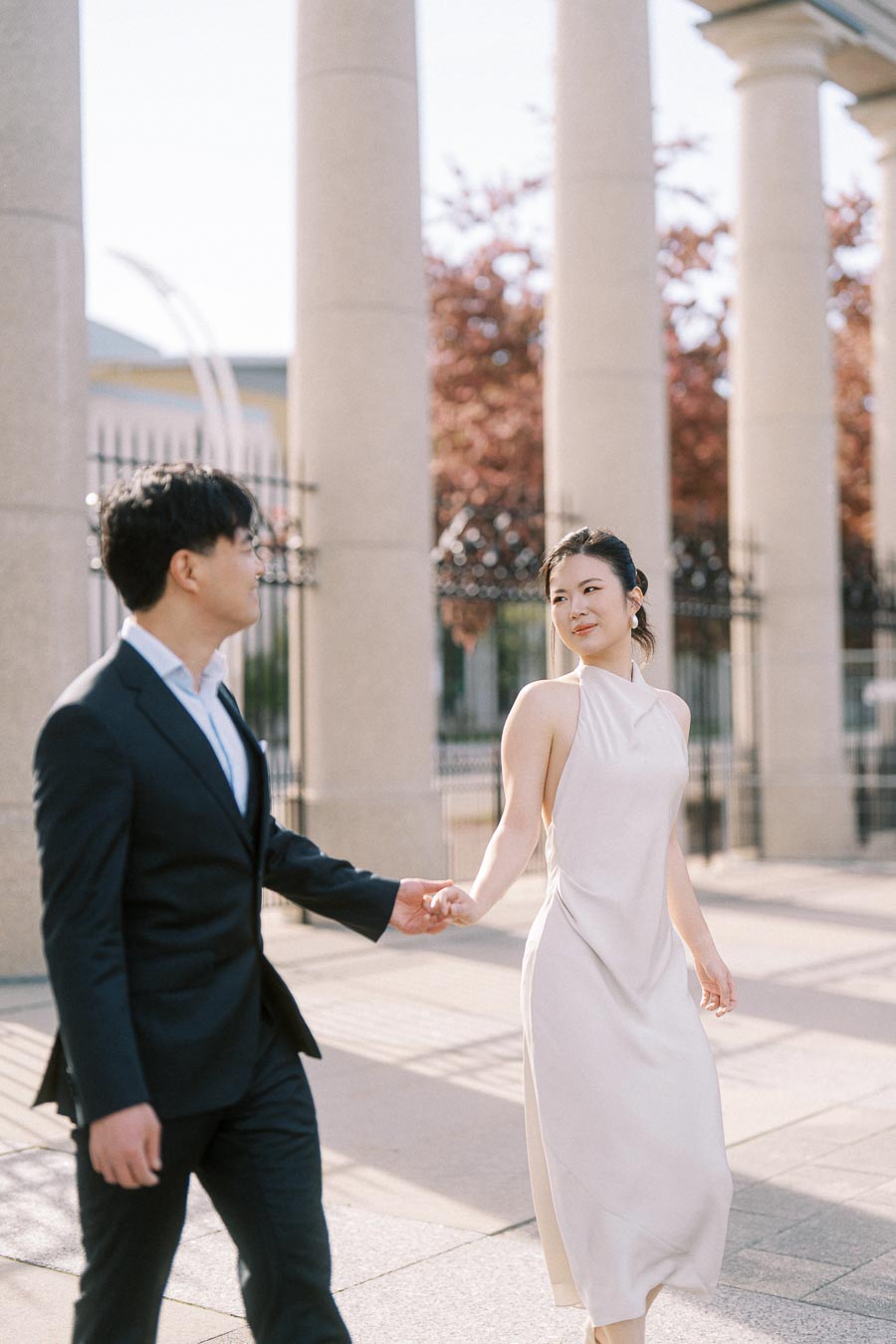 Engaged Asian couple walking hand in hand near the stone columns of the Princes Gates at Exhibition Place, Toronto, with soft sunlight and elegant attire.