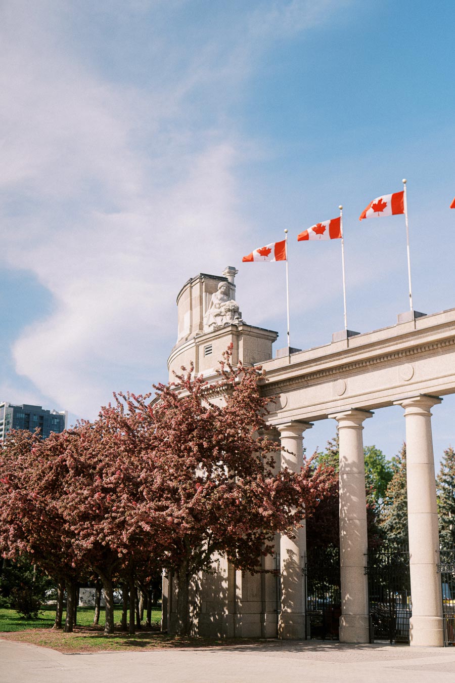 Princes' Gates at Exhibition Place in Toronto with pink flowering trees and Canadian flags flying above the stone columns on a sunny spring day.