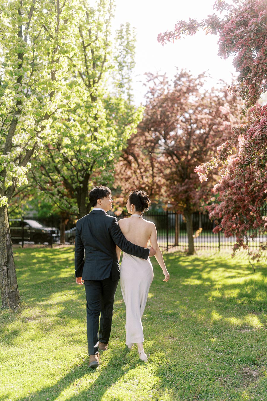Engaged couple walking arm in arm through Trillium Park in Toronto, surrounded by fresh spring greenery and pink flowering trees, backlit by soft sunlight.
