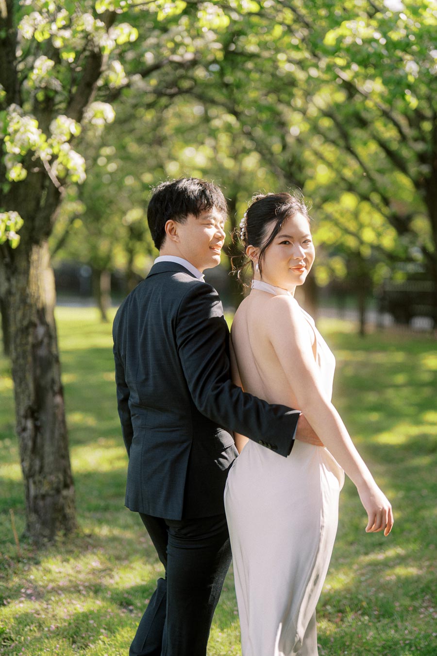 An engaged Asian couple stands closely together under spring trees at Trillium Park in Toronto, the woman in a white dress and the man in a black suit, bathed in soft sunlight.