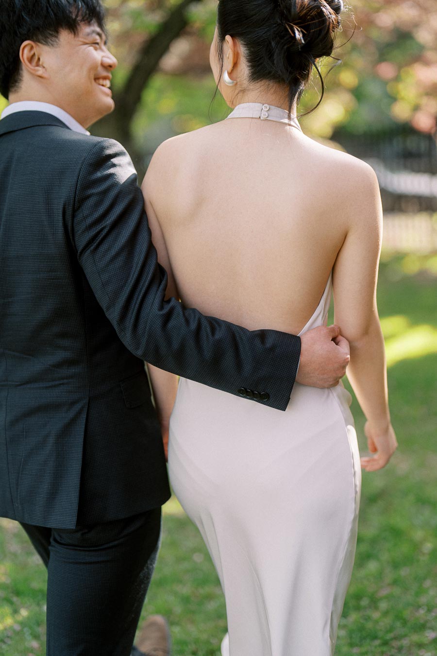 Engaged couple walking arm in arm at Trillium Park, Toronto. The groom-to-be smiles at his partner, who wears an elegant white dress, with soft sunlight and greenery in the background.