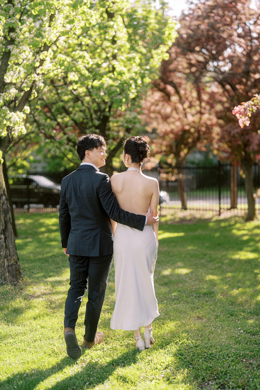 Engaged couple walking arm in arm through Trillium Park in Toronto, with spring trees and sunlight creating a romantic, elegant atmosphere.