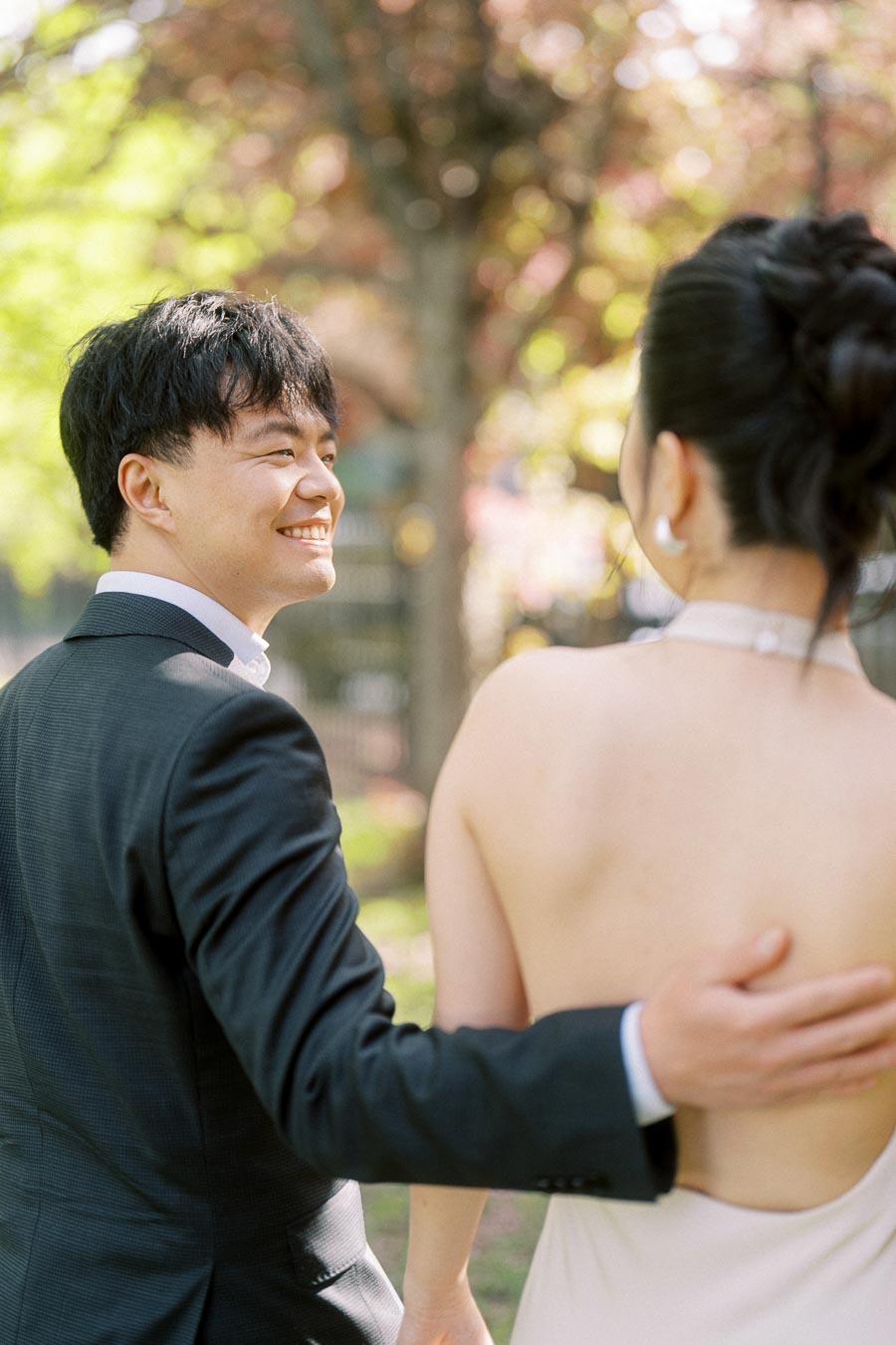 Asian engaged couple walking arm in arm in Trillium Park, Toronto, with the groom smiling warmly at the bride, surrounded by spring trees and sunlight.