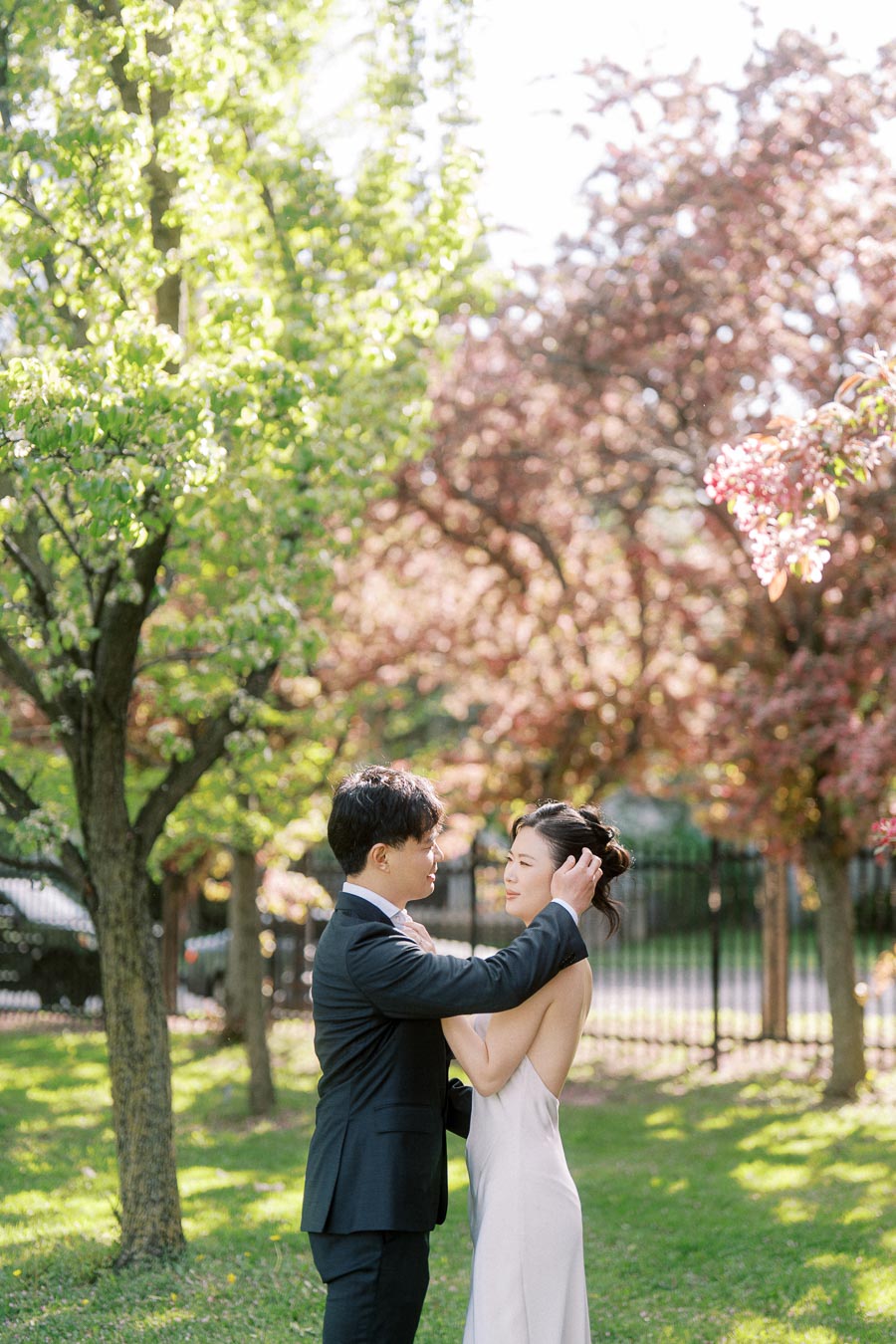 Engaged Asian couple sharing an intimate moment under blooming trees in Trillium Park, Toronto, with soft sunlight and spring greenery in the background.