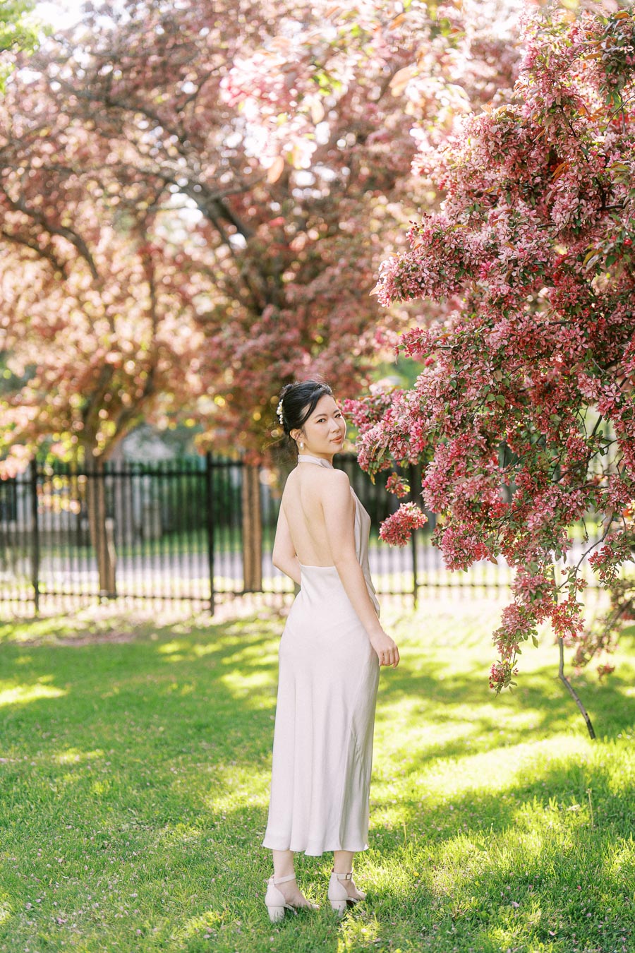 Asian bride-to-be in an elegant white dress posing beside blooming pink trees at Trillium Park, Toronto, with soft sunlight and spring greenery.