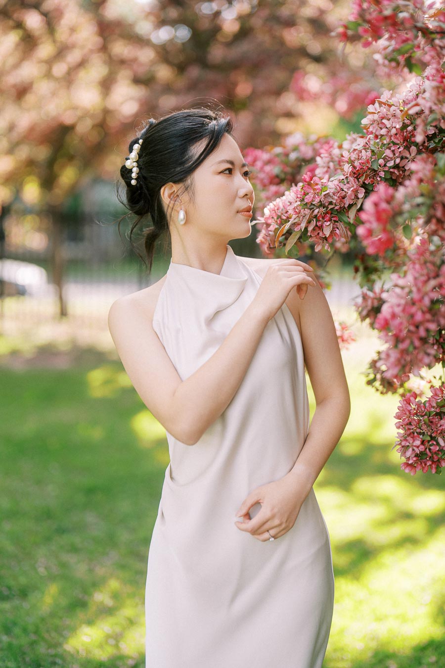 Asian bride to be in a white dress standing next to pink flowering tree branches at Trillium Park, Toronto, with soft sunlight and greenery in the background.