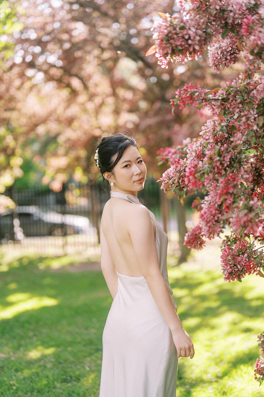 Asian bride-to-be in an elegant white dress stands beside vibrant pink flowering trees at Trillium Park, Toronto, with soft sunlight and greenery in the background.