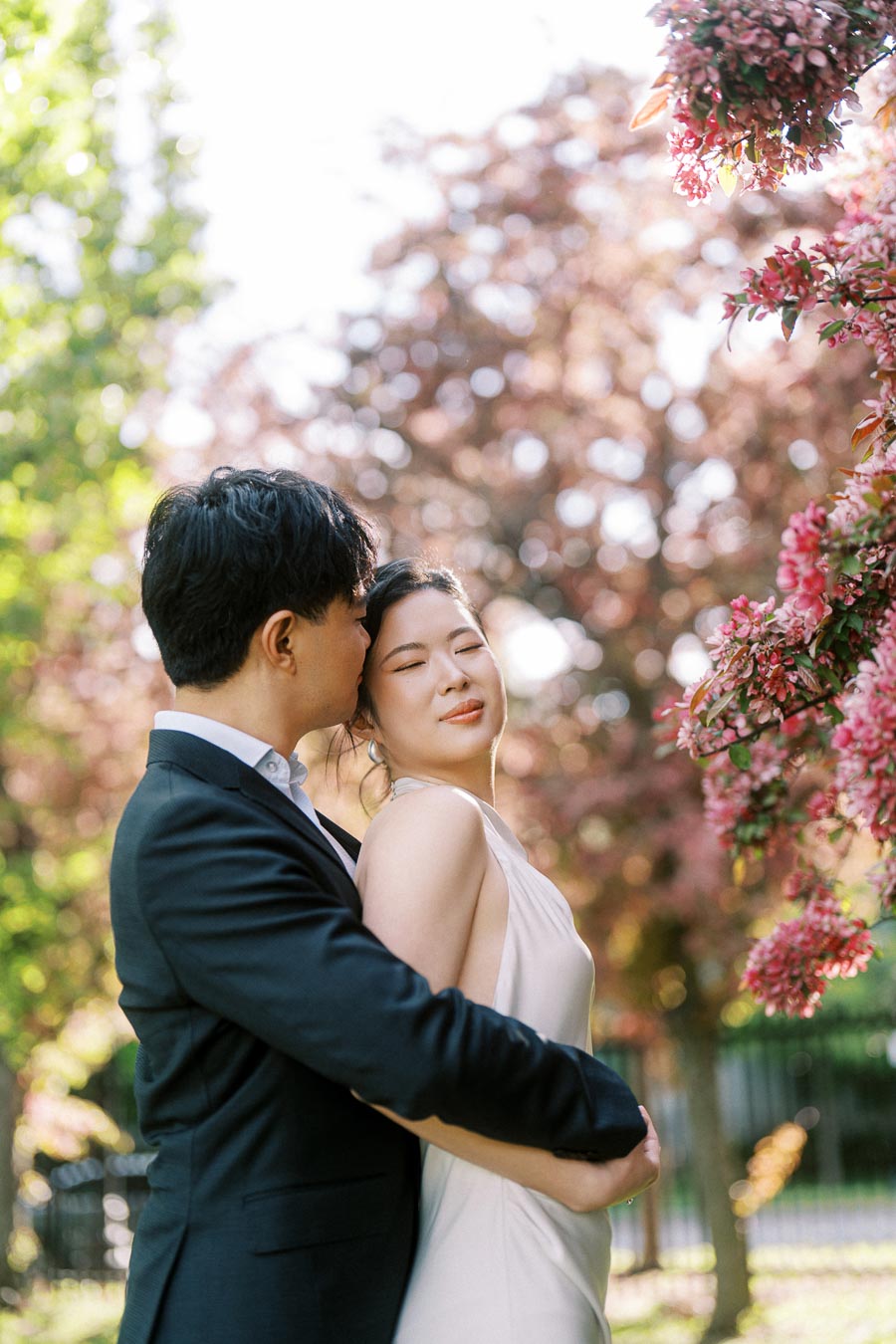 Asian engaged couple embracing under blooming pink trees in Trillium Park, Toronto, with soft sunlight and greenery in the background.