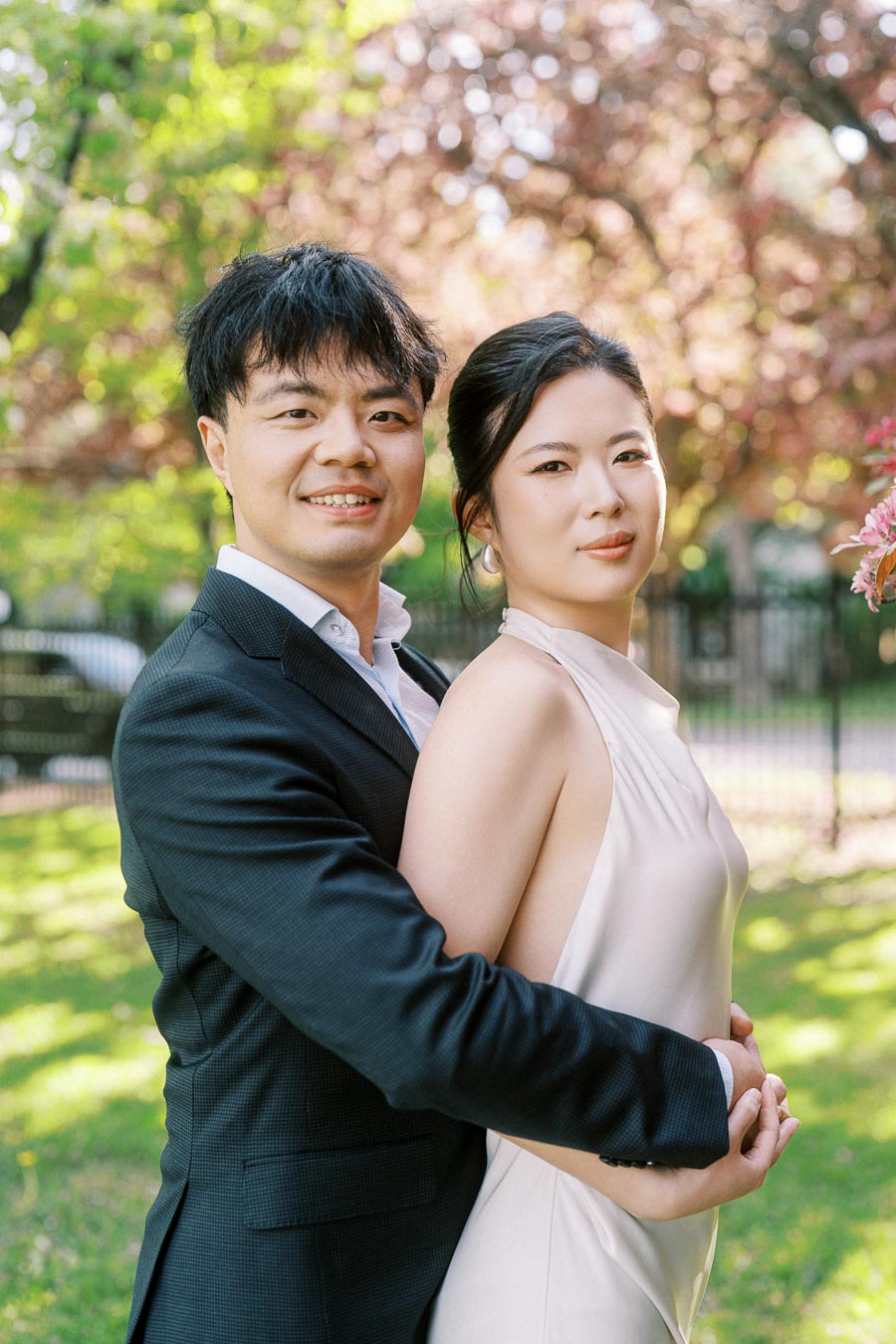 Engaged Asian couple standing close together in Trillium Park, Toronto, with the groom-to-be hugging the bride-to-be from behind amid spring trees and soft sunlight.