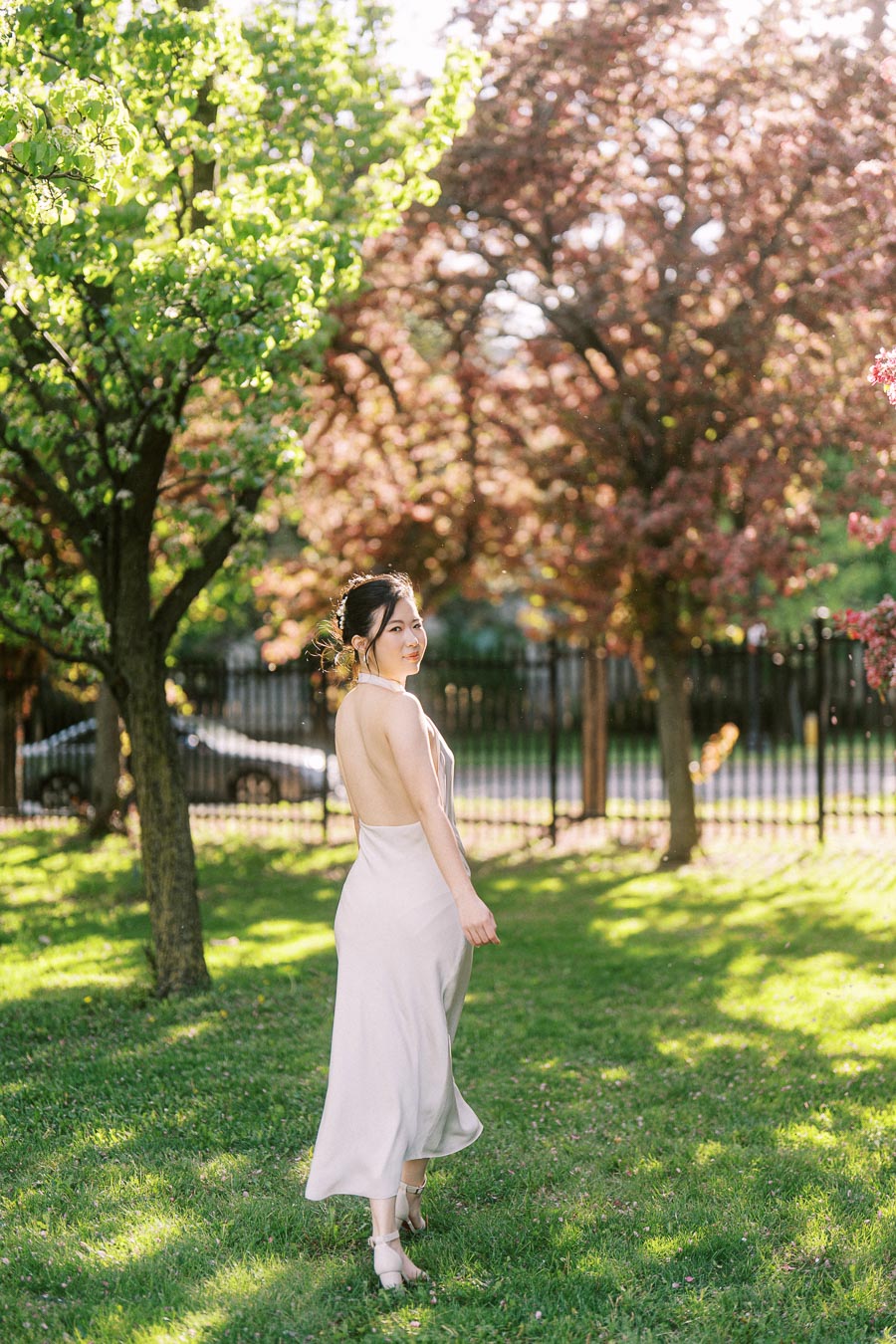 Bride-to-be in a backless white dress walking on green grass under blooming trees at Trillium Park, Toronto, in soft afternoon sunlight.