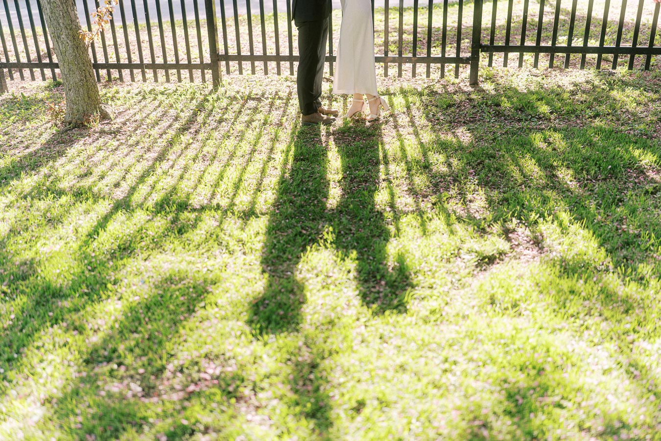 Artistic photo showing the lower halves of an engaged couple standing close together, with long shadows cast on sunlit grass and a black fence in Trillium Park, Toronto.
